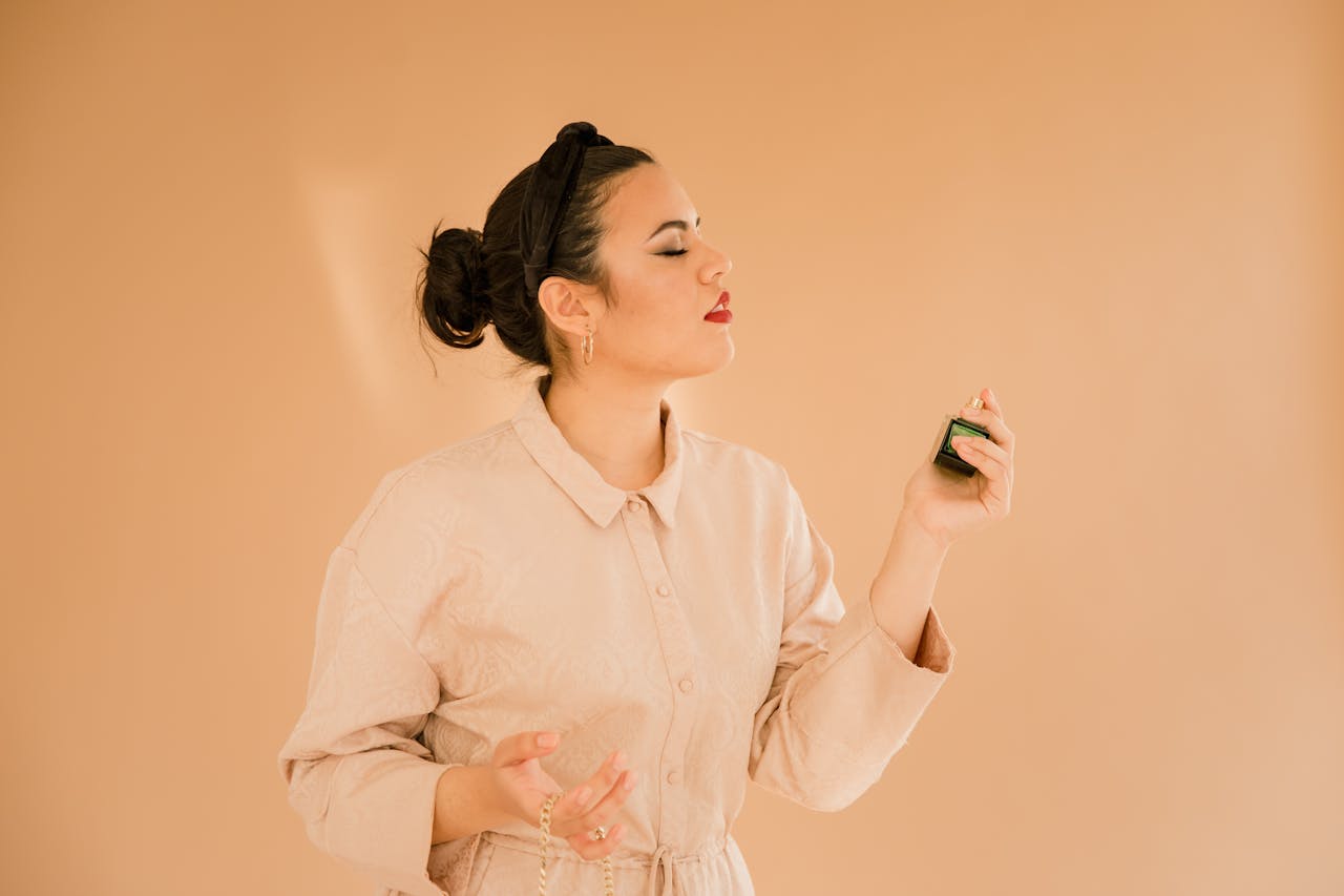 A woman in an elegant outfit applies perfume with grace against a neutral background.